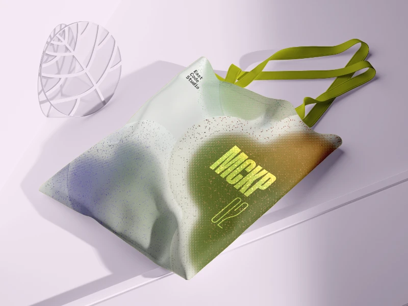 Eine Tragetasche mit einem Farbverlauf aus Grün, Weiß und Violett und einer gesprenkelten Textur wird auf einer hellvioletten Fläche präsentiert. Die Tasche hat zwei grüne Träger, die über sie drapiert sind. Links neben der Tasche befindet sich ein durchscheinendes, blattförmiges Objekt mit einem geometrischen Muster. Der Hintergrund ist einfarbig hellviolett mit weichen Schatten. Die Gesamtkomposition ist klar und minimalistisch, wobei der Fokus auf der Tragetasche liegt.