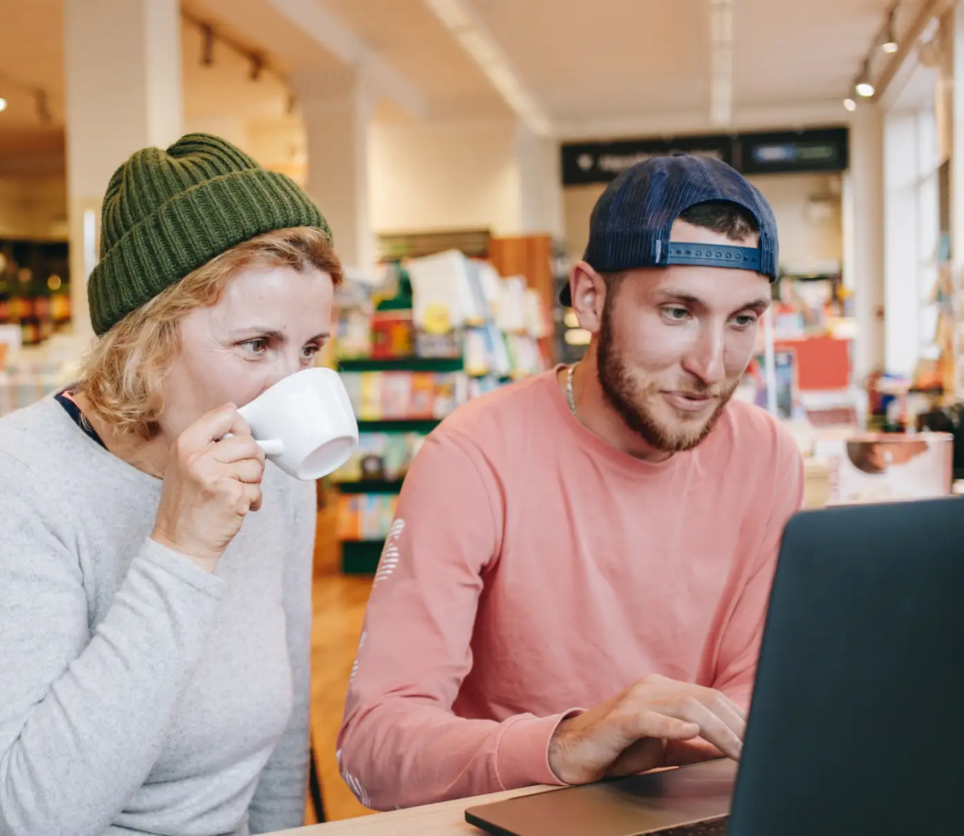 Eine Nahaufnahme zeigt eine Frau und einen Mann, die an einem Tisch in einer hell erleuchteten Buchhandlung sitzen. Die Frau auf der linken Seite trägt einen grauen Pullover und eine grüne Mütze und trinkt aus einer weißen Tasse. Der Mann auf der rechten Seite trägt ein rosa Langarmshirt und eine blaue Baseballkappe und schaut auf einen Laptop. Der Hintergrund ist unscharf und zeigt Bücherregale voller Bücher und andere Kunden, die stöbern. Die allgemeine Stimmung ist ungezwungen und entspannt.